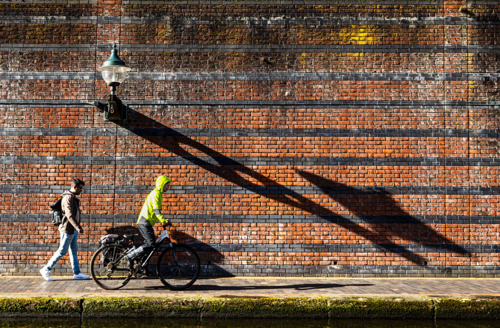 A cyclist and a pedestrian approach a long shadow of a street lamp.